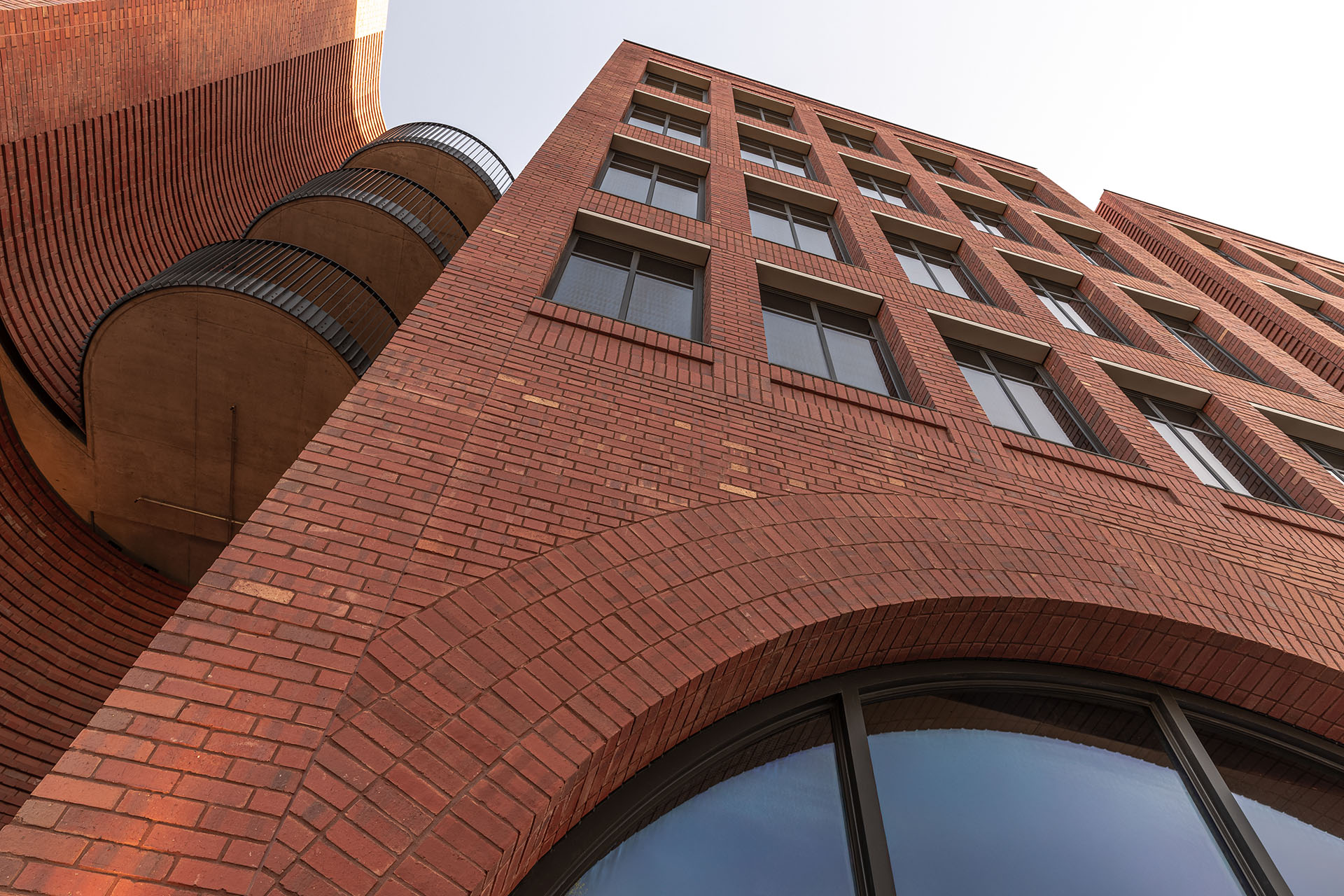 Looking up a red brick building facade, curved brickwork and rows of windows against a pale sky